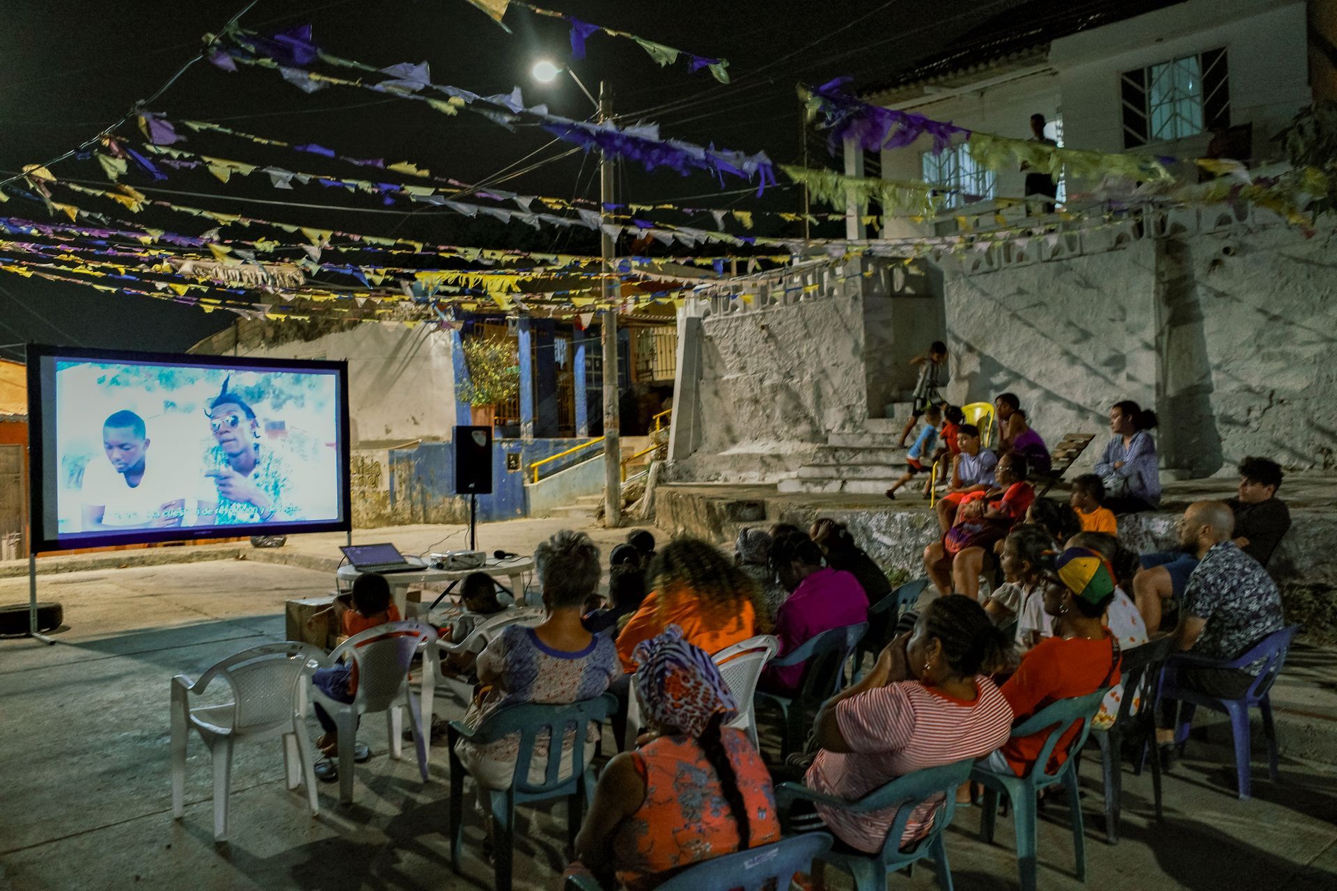 Proyección de cine al aire libre con público sentado bajo luces de guirnalda por la noche.
