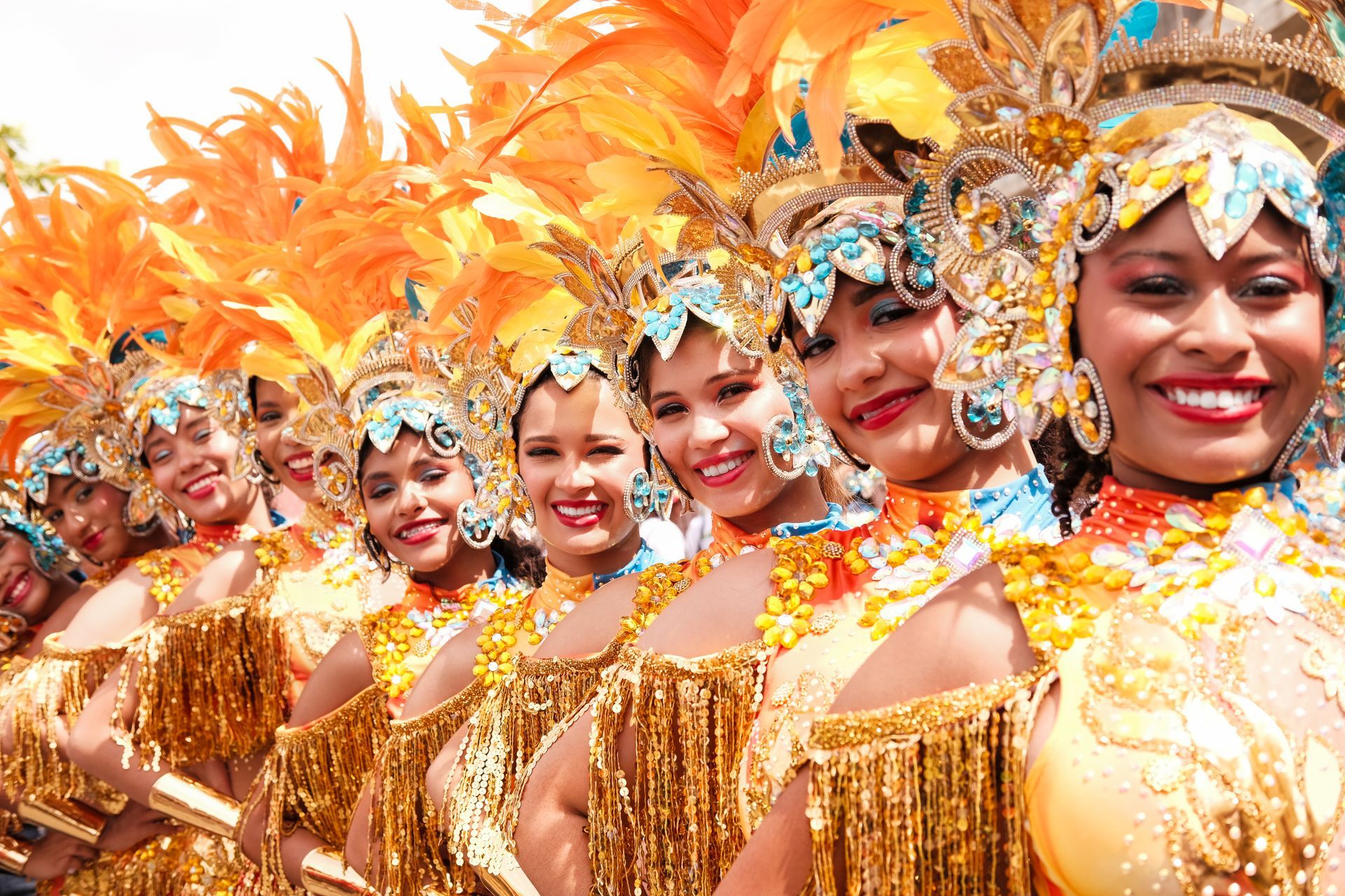 Un colorido desfile de bailarines con trajes dorados y tocados de plumas naranjas, sonriendo en una formación festiva.