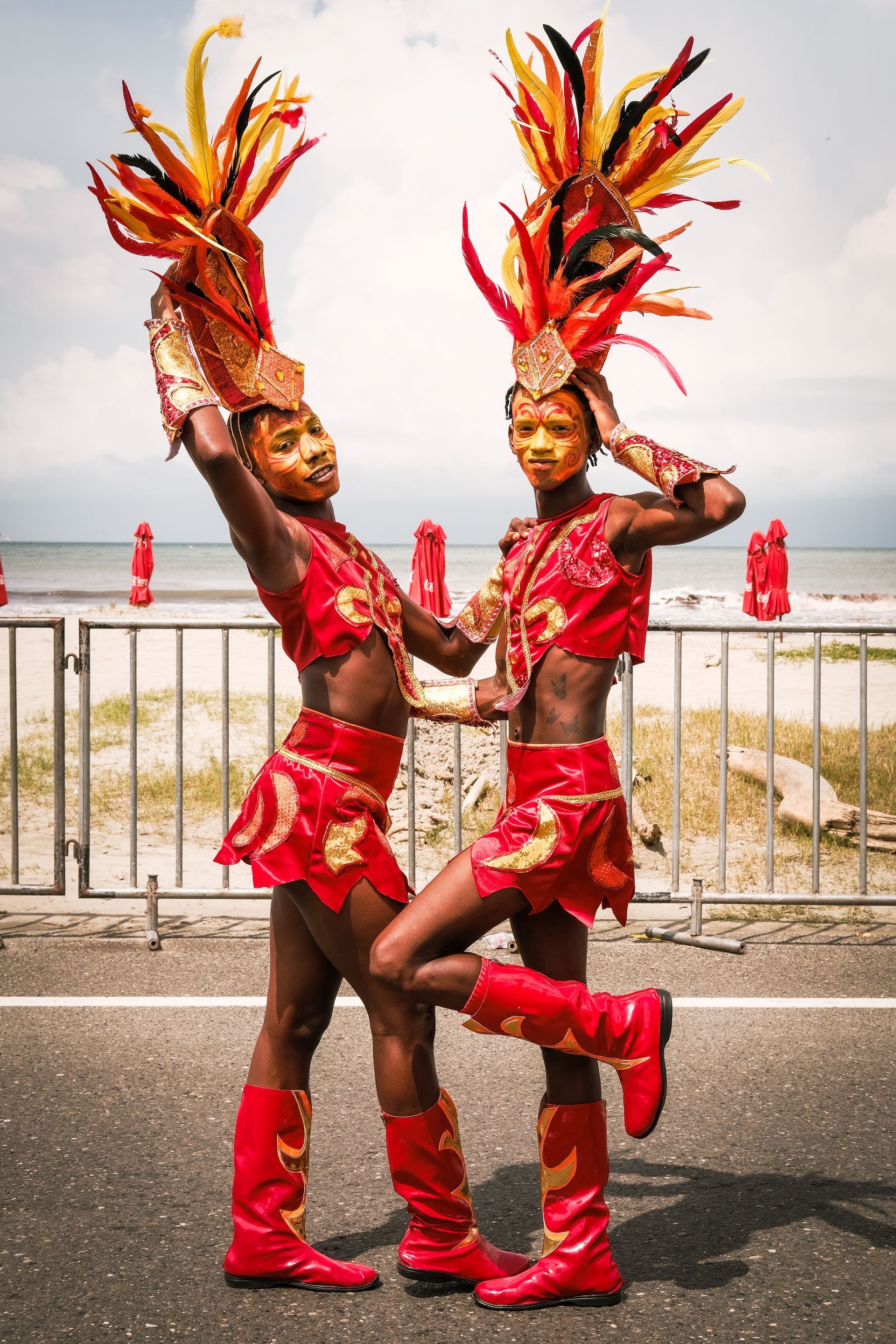 Dos artistas con trajes rojos y tocados de plumas posan al aire libre cerca de una valla junto al paseo marítimo.