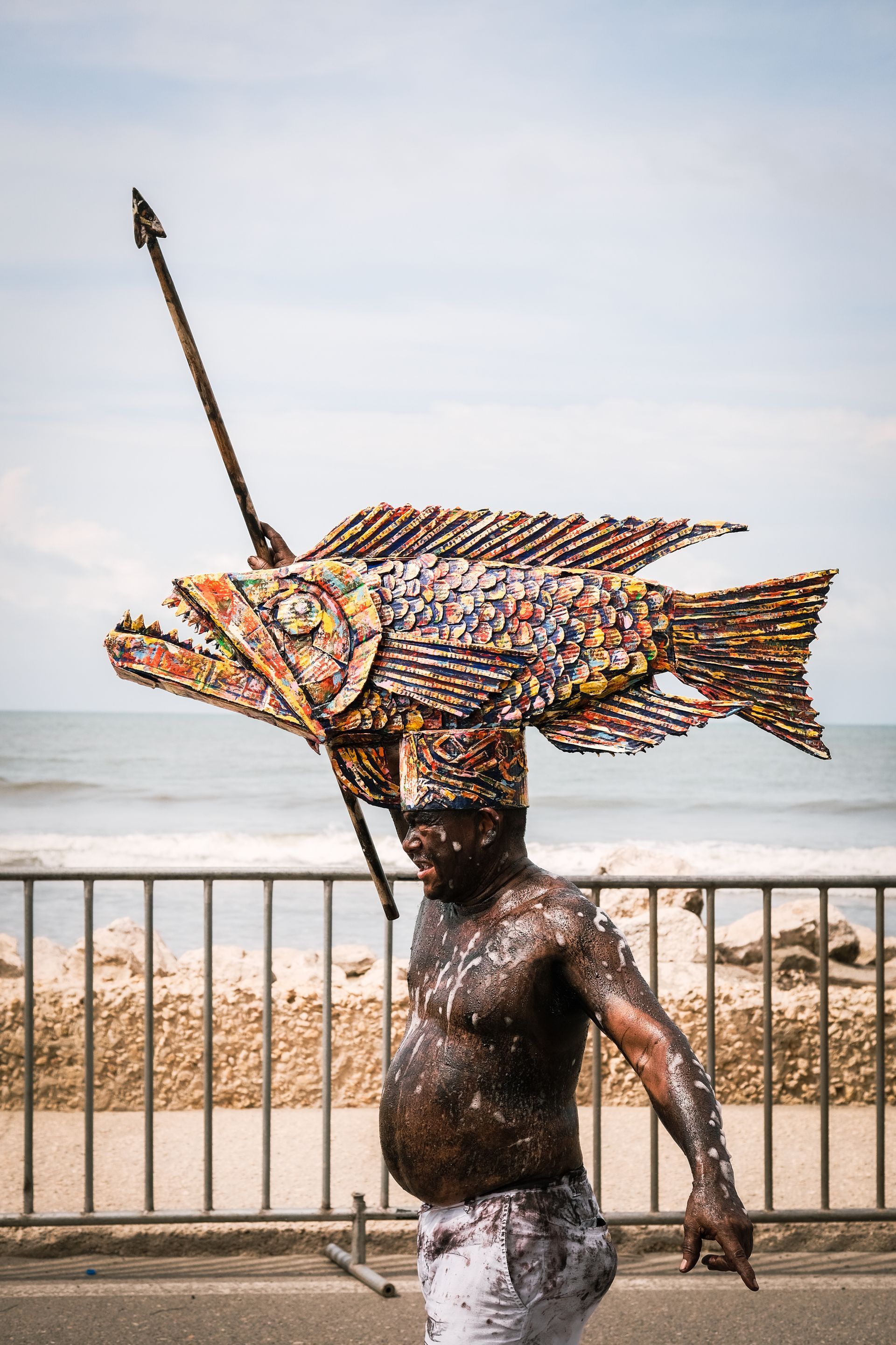 Una persona lleva una colorida escultura de un pez sobre la cabeza junto a la barandilla de una playa.