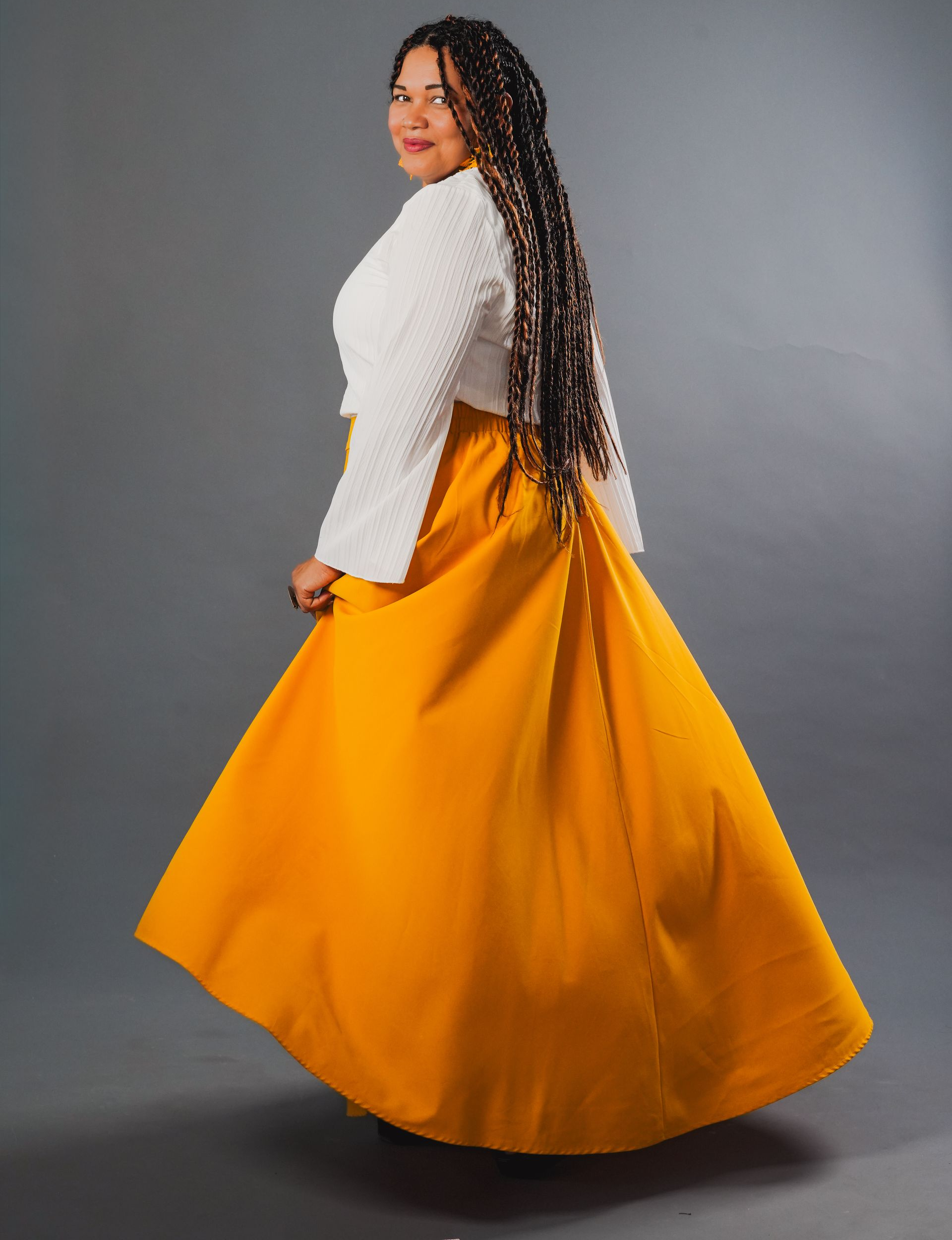 Persona con una blusa blanca y una falda fluida de color amarillo mostaza posando frente a un fondo gris.