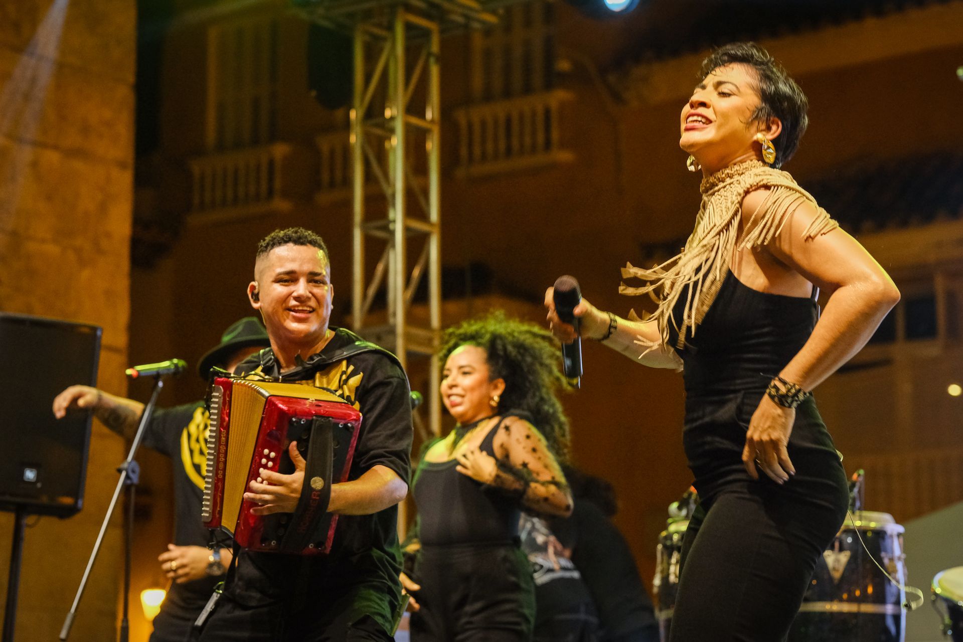 Tres músicos actuando en el escenario, con acordeón, micrófono y movimientos de baile bajo luces cálidas.