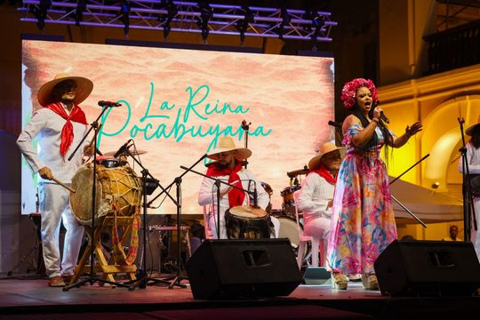 Banda en vivo actuando en el escenario con una cantante con un vestido colorido y percusionistas bajo una iluminación cálida.