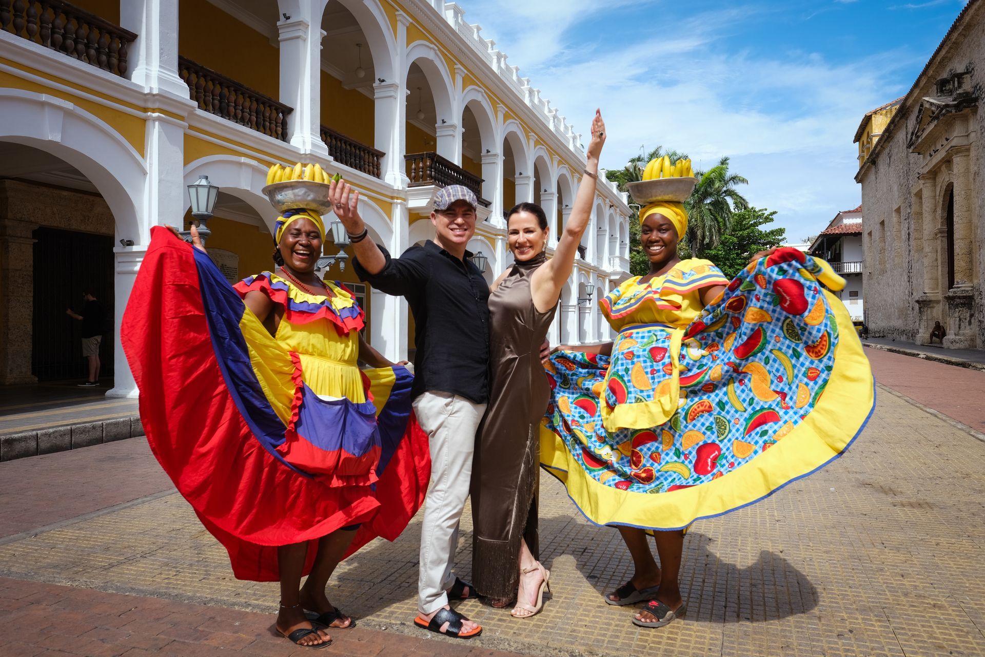 Tres bailarinas posando con coloridos trajes tradicionales en una calle empedrada junto a un edificio histórico.