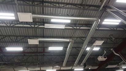 Metal HVAC ductwork and rectangular ceiling vents suspended from a black industrial ceiling with linear light fixtures.