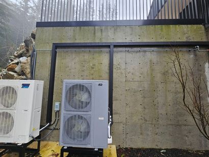 Two HVAC units sit on black stands against a concrete wall with a metal railing above and a rock feature on the left.