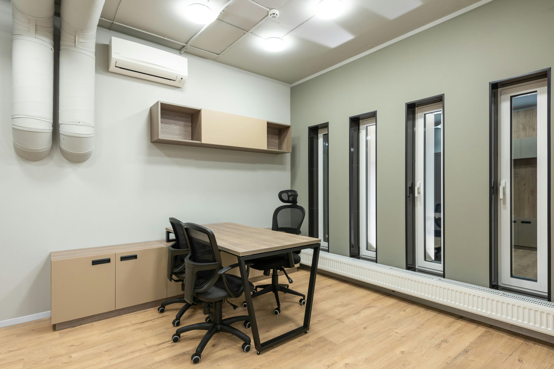 A modern office meeting room with a wooden table, three black office chairs, wall shelving, and tall windows on green walls.