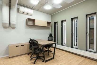 A modern office meeting room with a wooden table, three black office chairs, wall shelving, and tall windows on green walls.