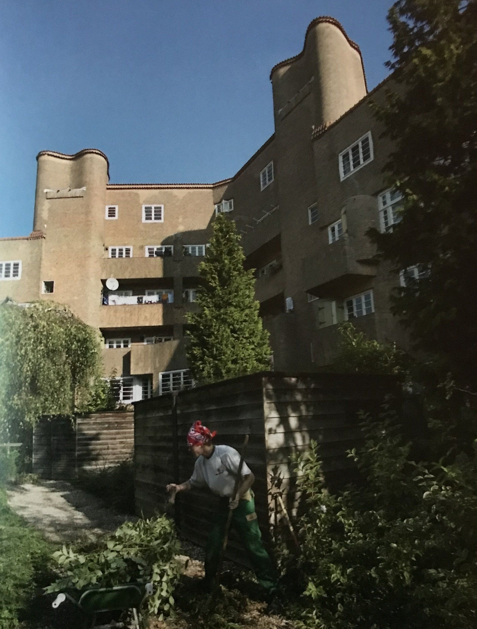 Een persoon is aan het tuinieren voor een beige appartementencomplex met ronde torens. Blauwe lucht.