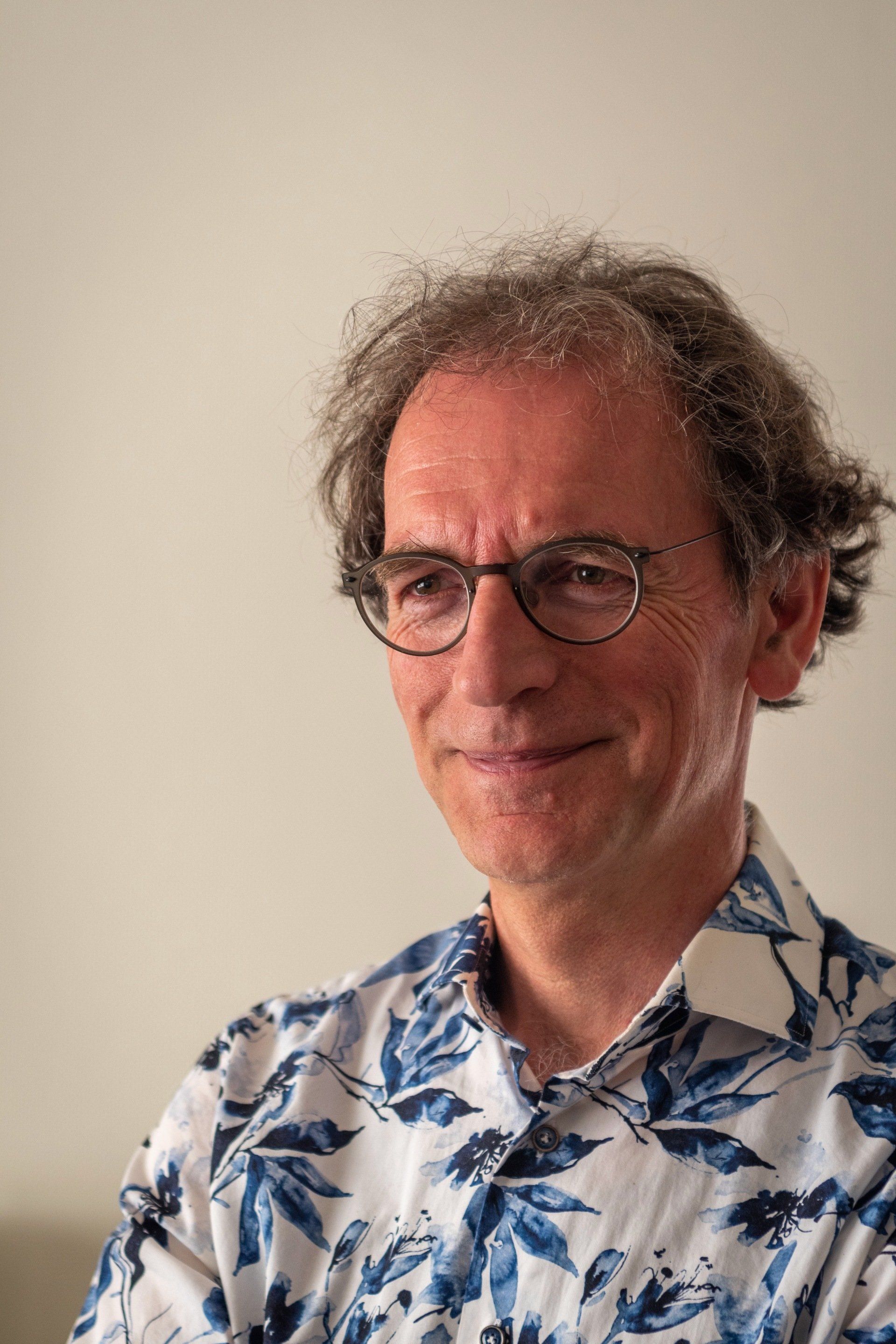 Een man met een bril en krullend haar lacht in een wit-blauw bloemenhemd.