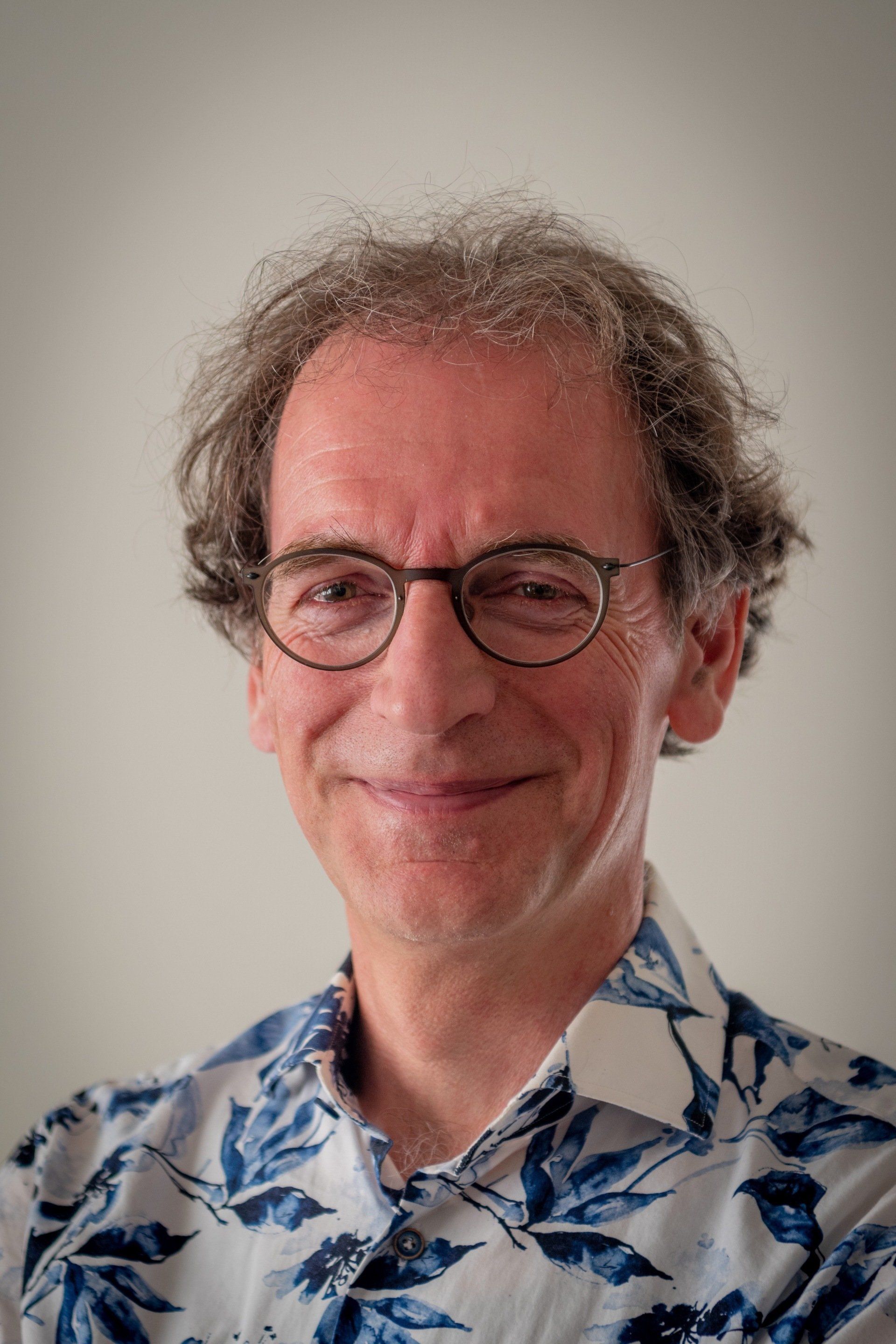 Man met bril, gekleed in een wit-blauw bloemenhemd, lacht naar de camera.