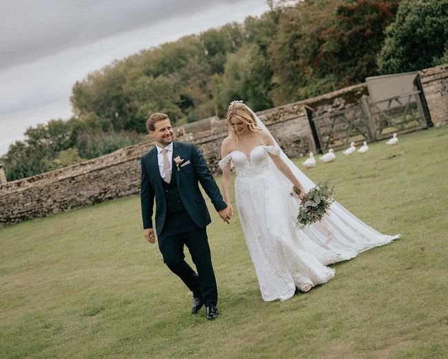 A bride and groom are walking in a field holding hands.