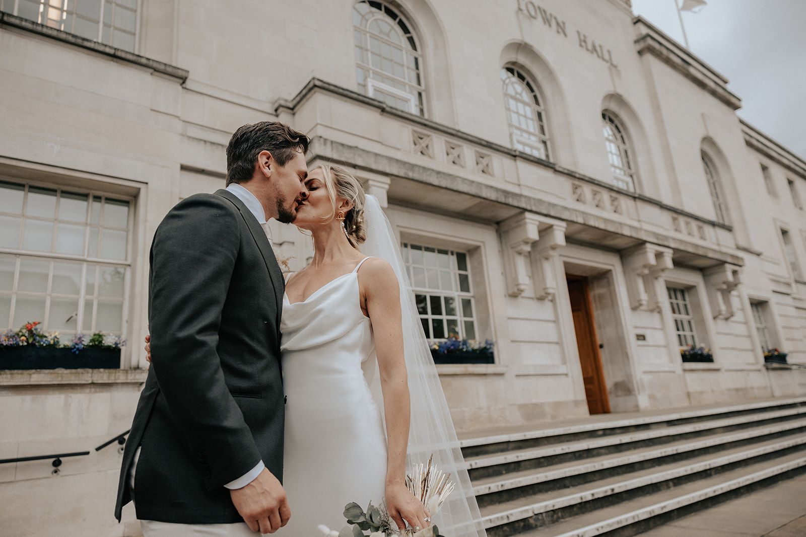 A black and white photo of a bride and groom kissing in front of a brick building.