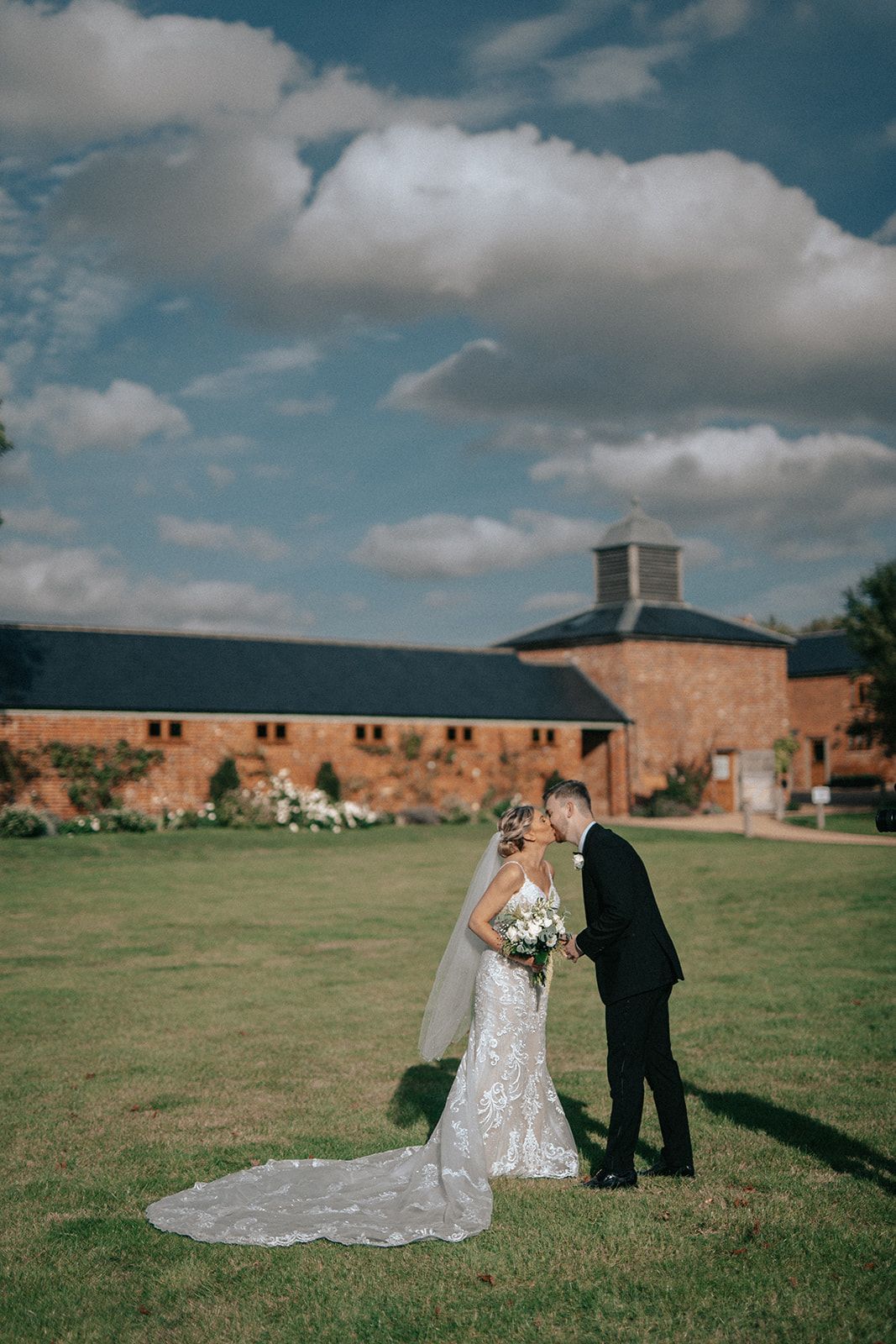 A bride and groom are kissing in a field in front of a building.