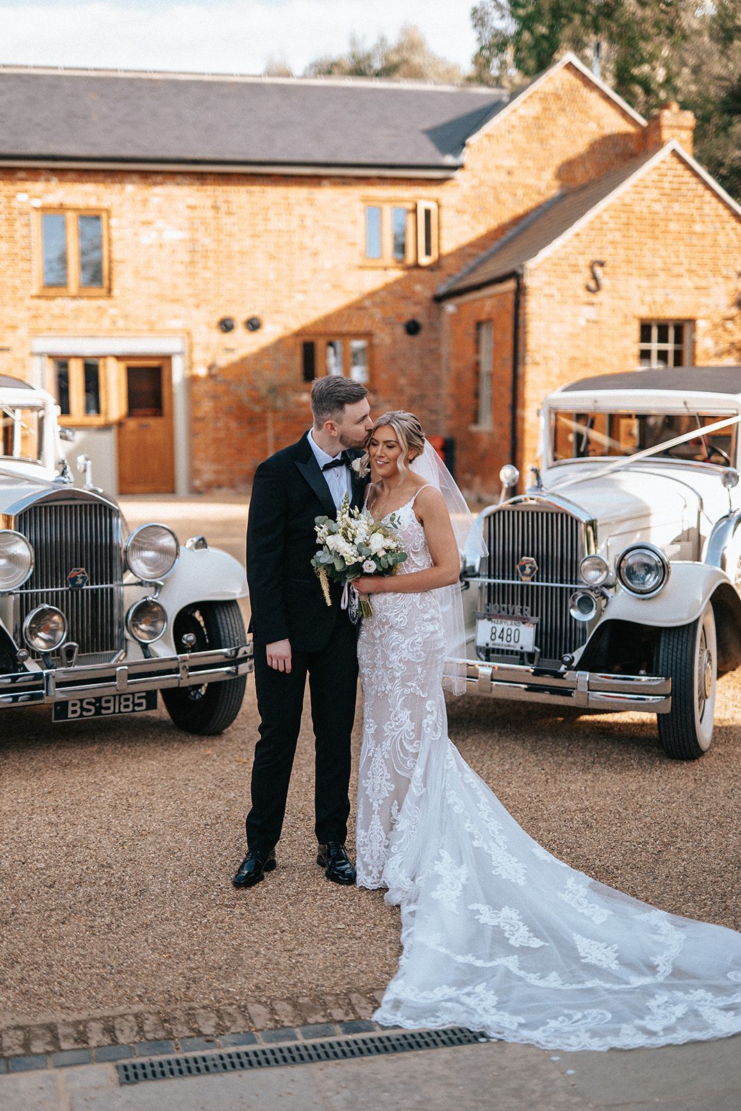A bride and groom are kissing in front of two vintage wedding cars.
