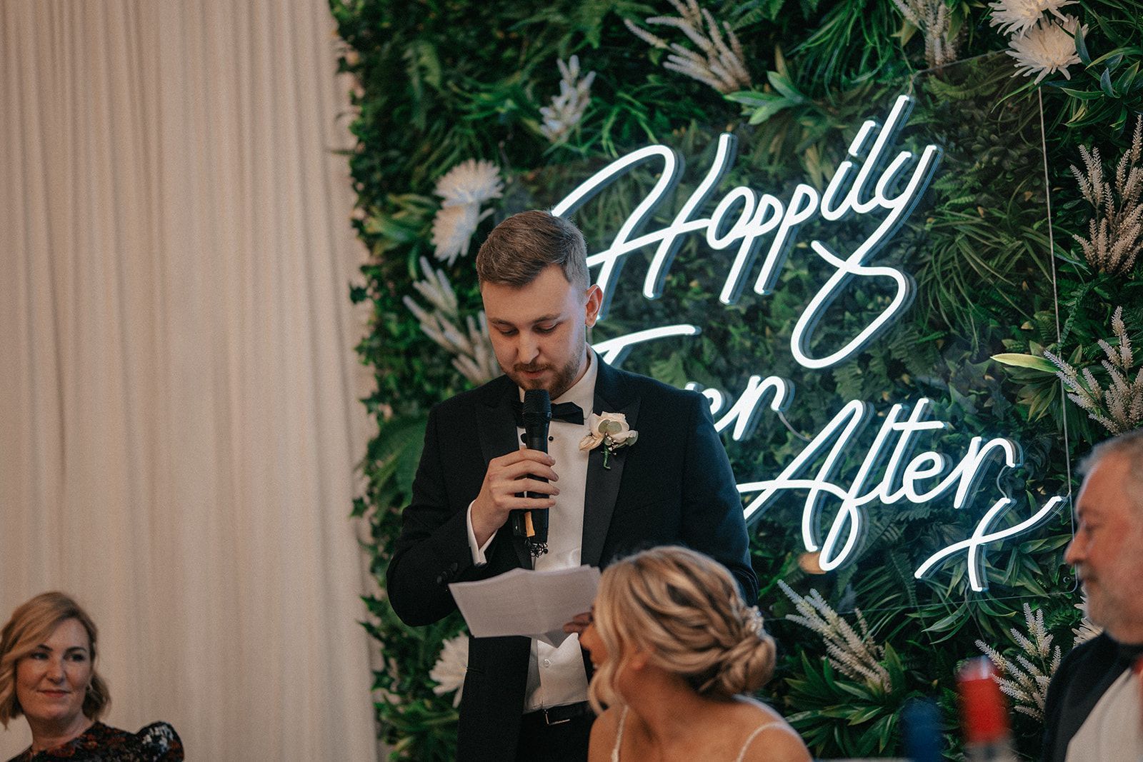 A man is giving a speech at a wedding reception in front of a neon sign that says `` happily ever after ''.