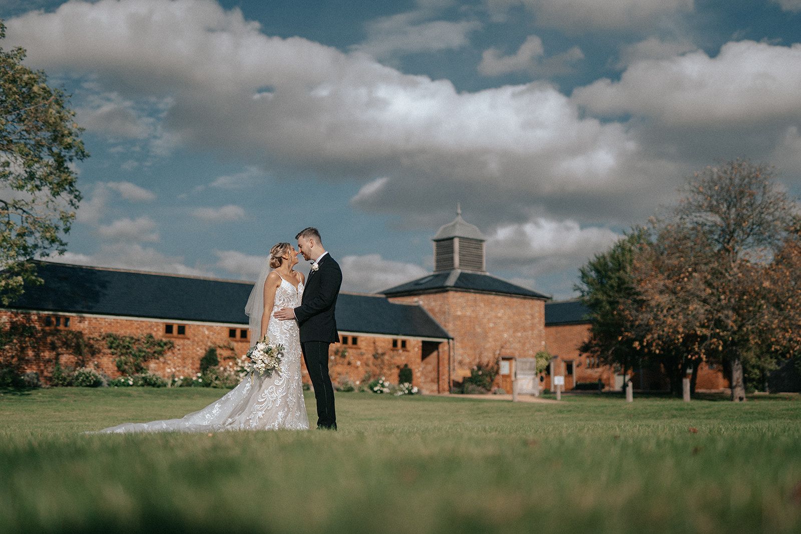A bride and groom are standing in a field in front of a brick building.
