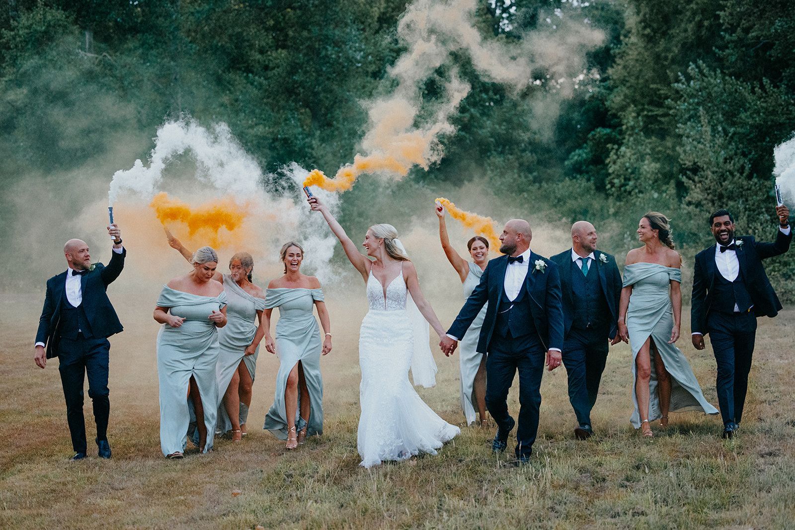 A bride and groom are walking through a field with their wedding party holding smoke grenades.