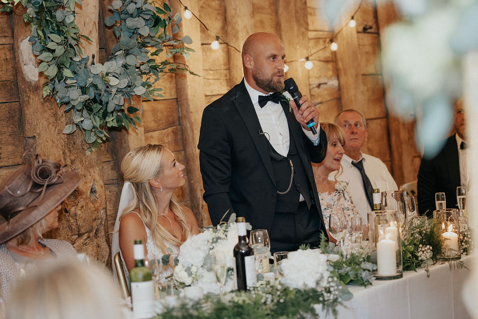 A man in a tuxedo is giving a speech at a wedding reception.