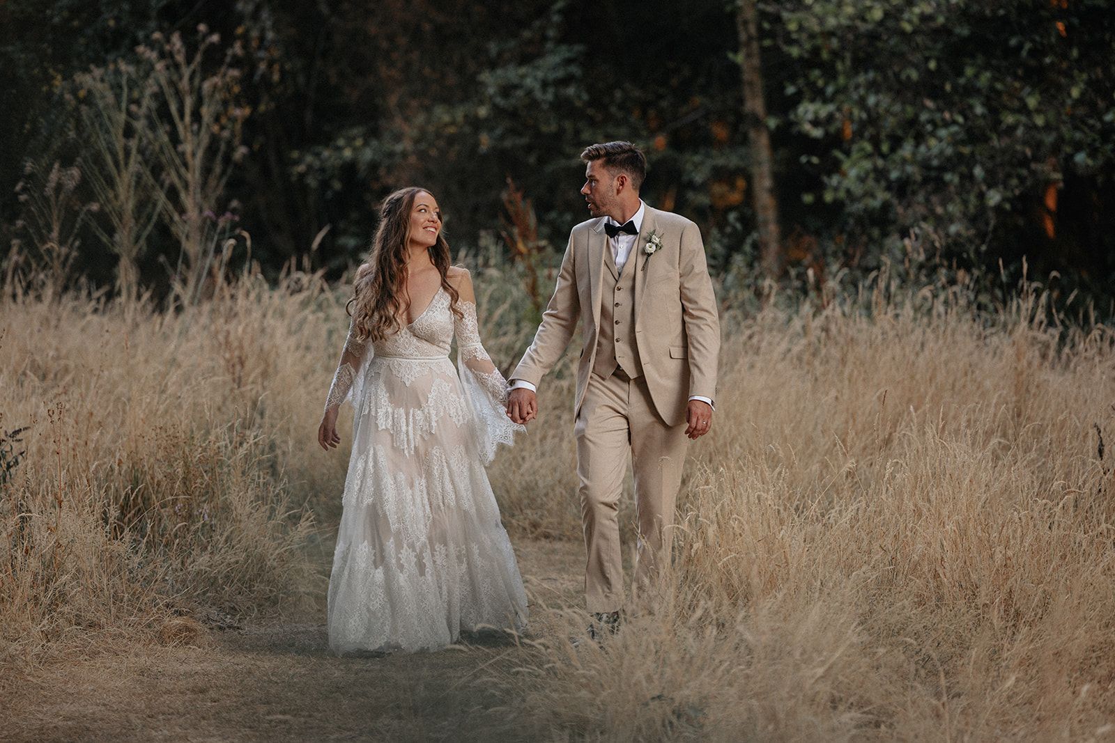 A bride and groom are walking through a field holding hands.