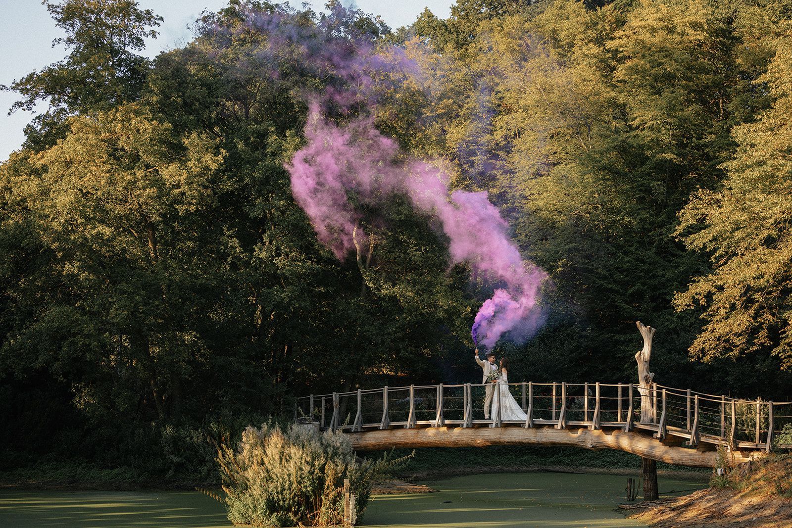 A bride and groom are standing on a bridge with purple smoke coming out of it.
