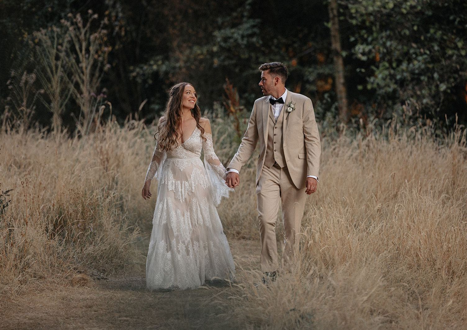 A bride and groom are walking through a field holding hands.