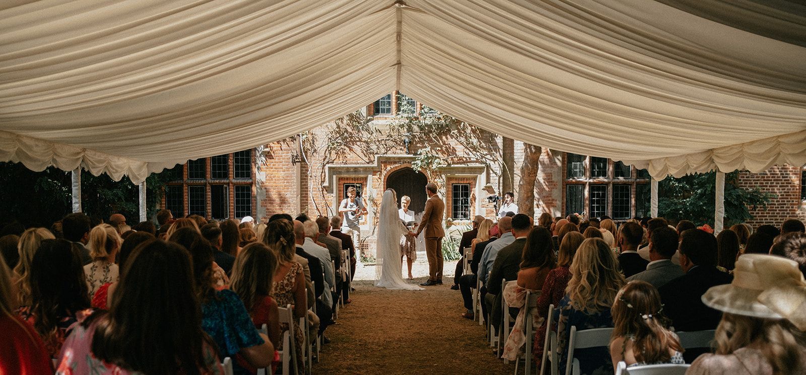 A large group of people are sitting under a tent watching a wedding ceremony.