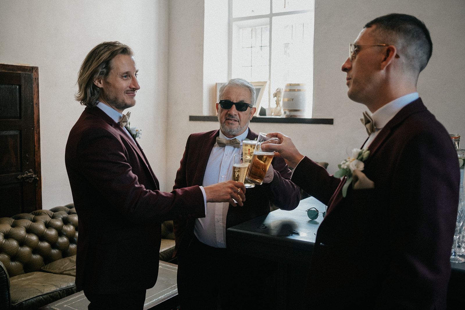 Three men in suits are toasting with beer in a room.