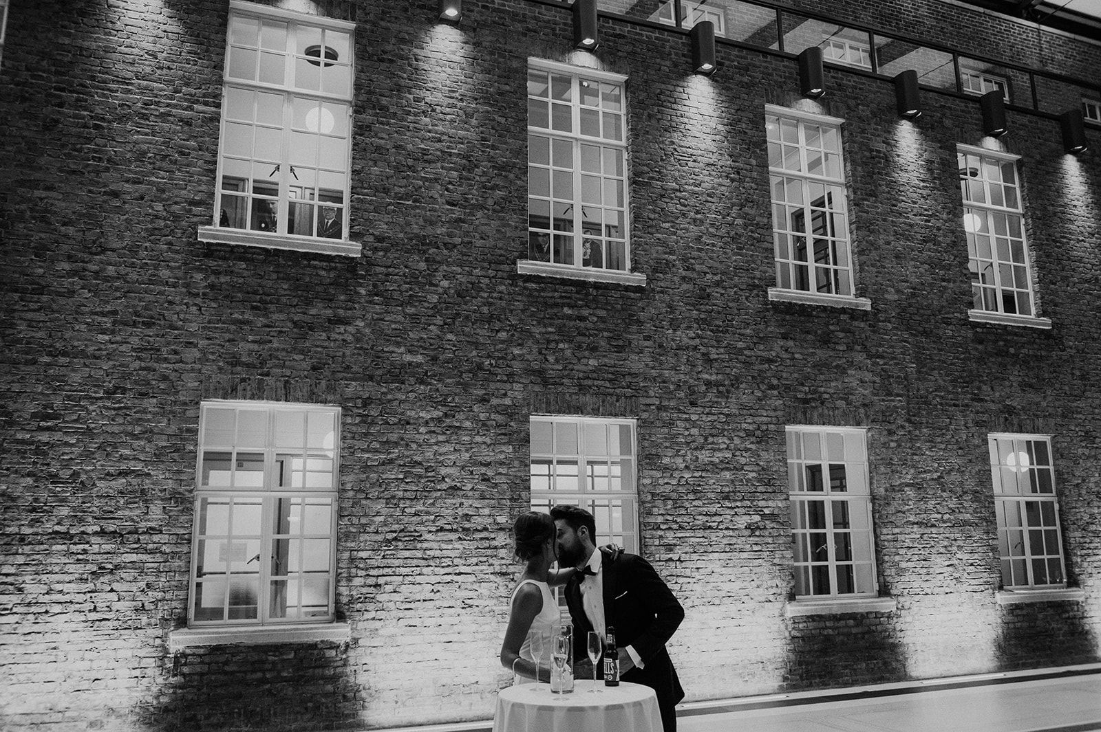 A black and white photo of a bride and groom kissing in front of a brick building.