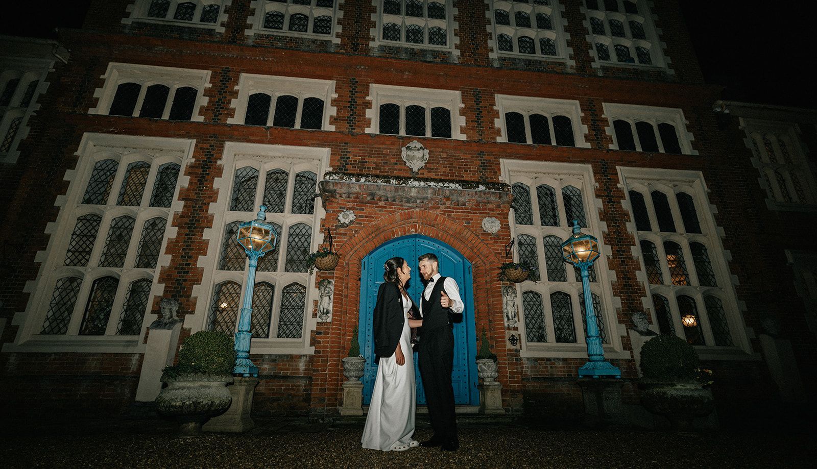 A bride and groom are standing in front of a large brick building at night.