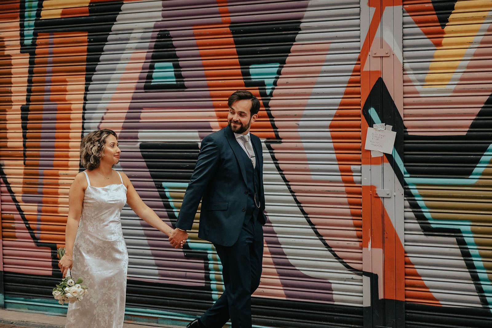 A bride and groom are holding hands in front of a graffiti wall.