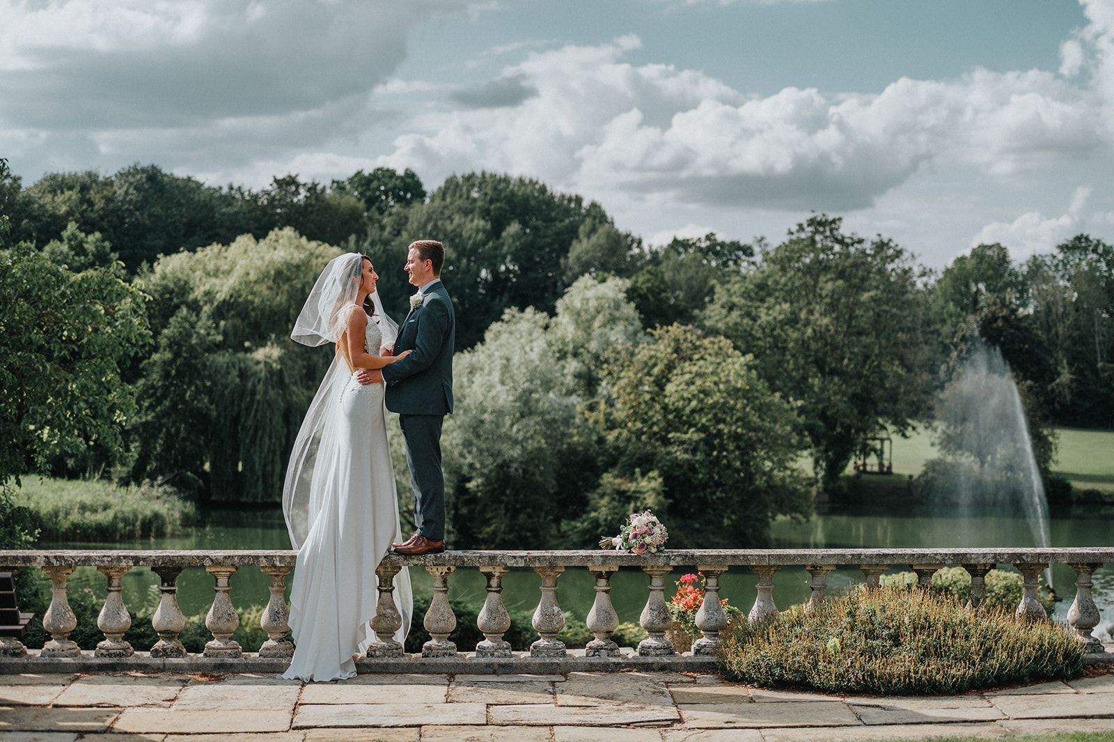 A bride and groom are standing on a balcony overlooking a lake.