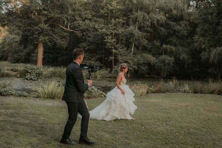 A man is holding a camera while a bride in a wedding dress is walking in the grass.