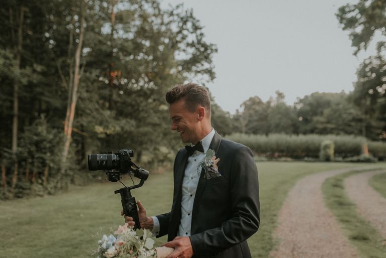a man in a tuxedo is holding a camera and a bouquet of flowers .