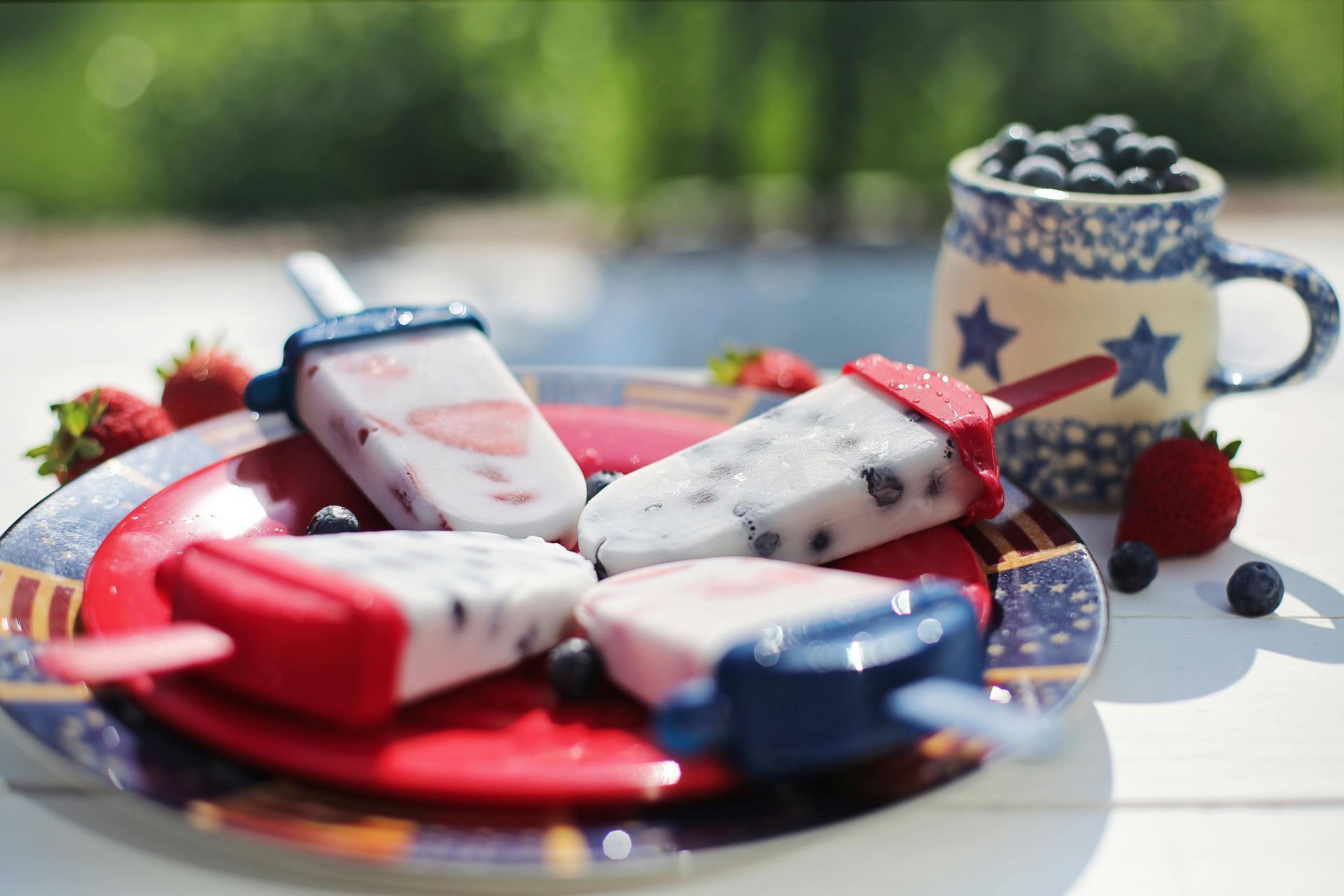 Red, white, and blue popsicles on a plate with blueberries, strawberries, and a star-spangled mug.