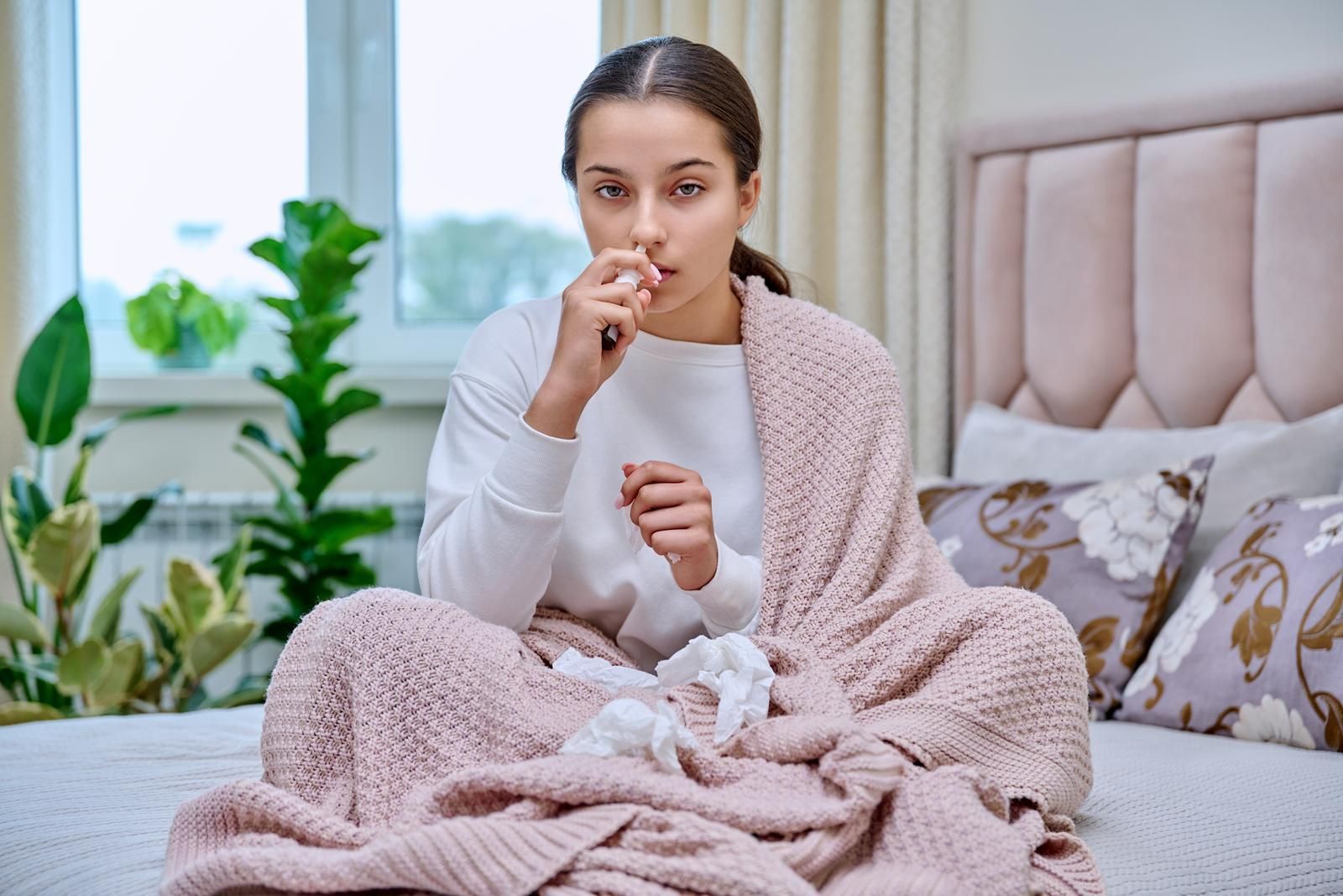 A woman using nasal moisturizer and is sitting on a bed wrapped in a blanket.