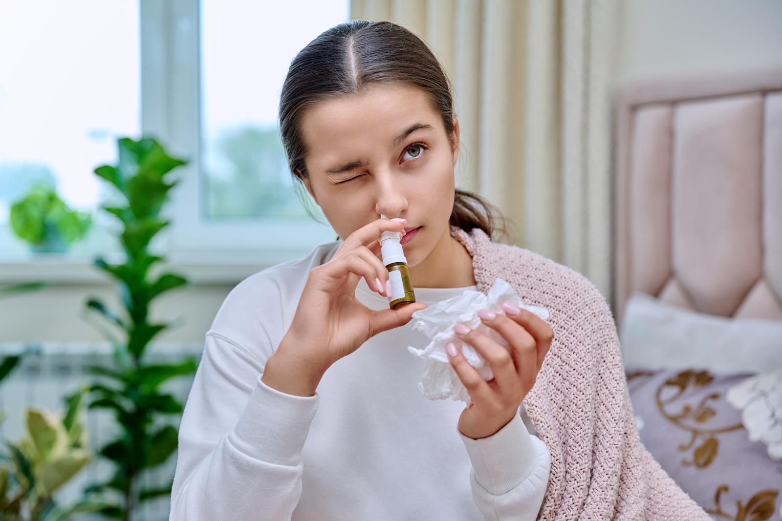 A young woman is sitting on a bed using a nasal gel/spray.