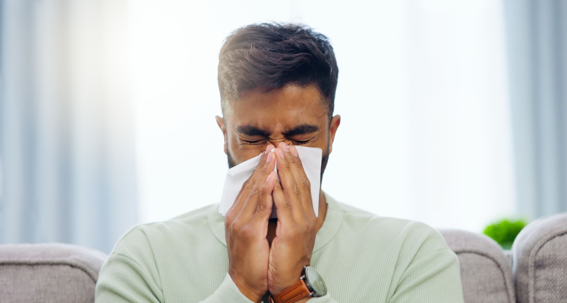 A man suffering from dry sinuses is blowing his nose into a napkin while sitting on a couch