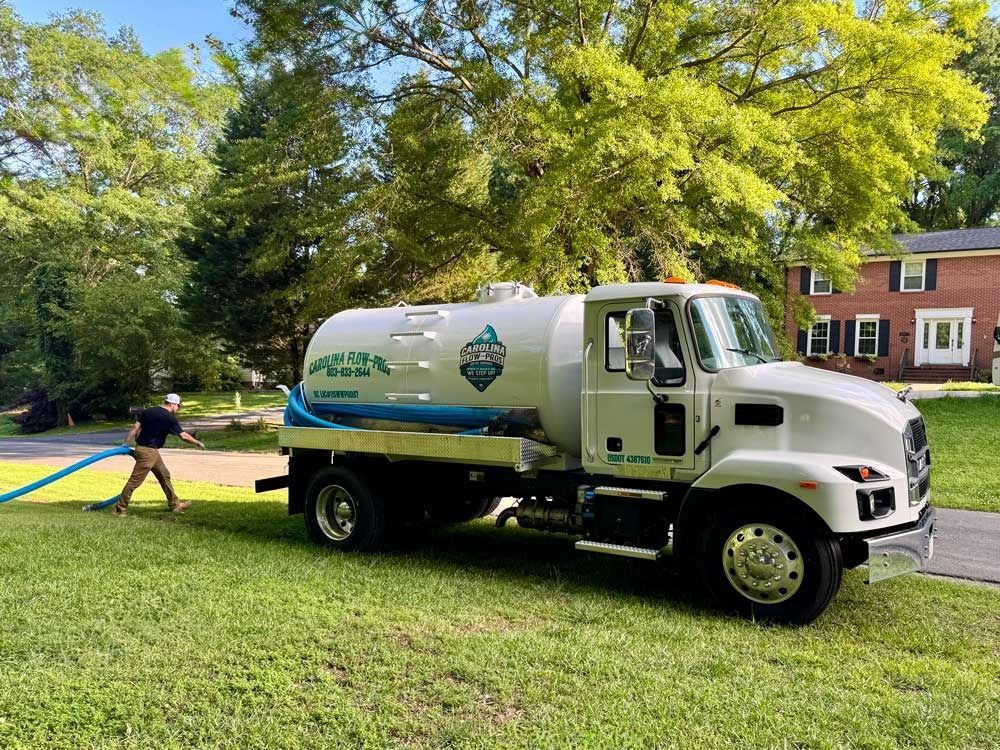 White septic tank truck on grass; man holding blue hose, house in background.