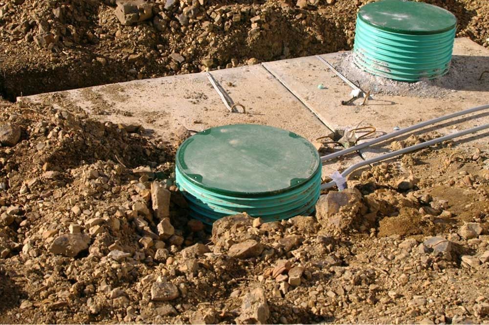 Two green septic tank covers in dirt, near a concrete pad with pipes.
