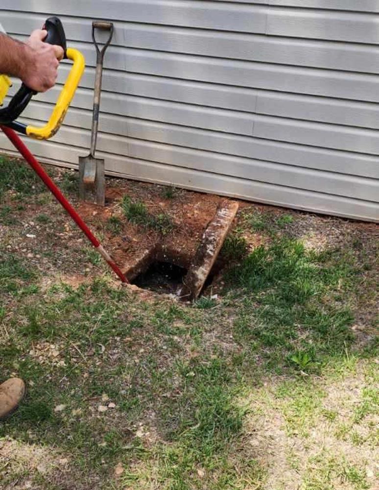 Person using tool near open access point in yard beside a building.