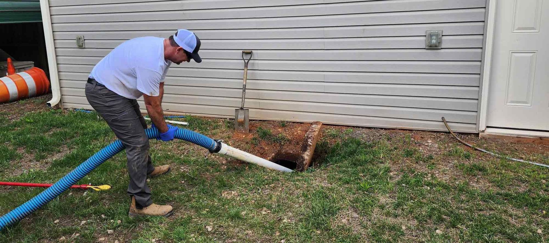 A man in a baseball cap is pumping a septic tank in front of a white building.
