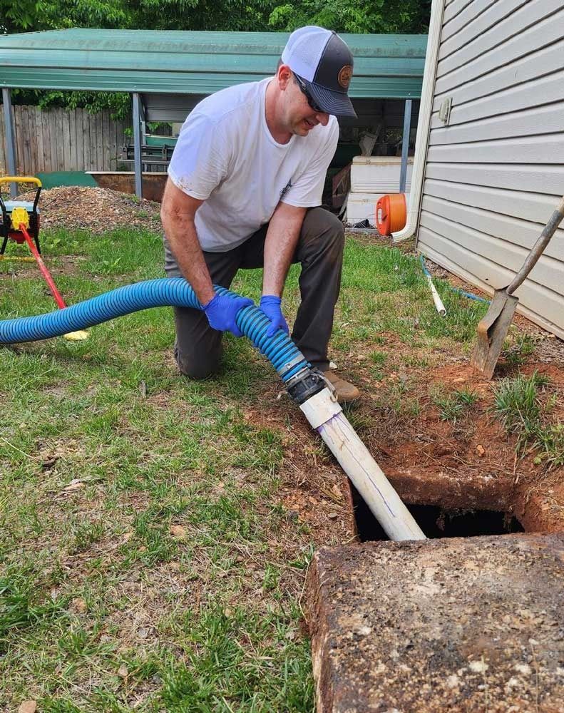 Man in white shirt kneels, holding hose into open septic tank in a yard.