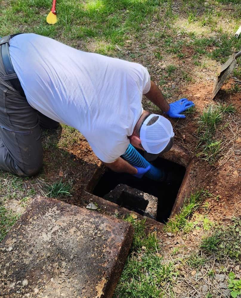A person wearing gloves is inspecting a septic tank. They are kneeling, looking down into an open square hole.