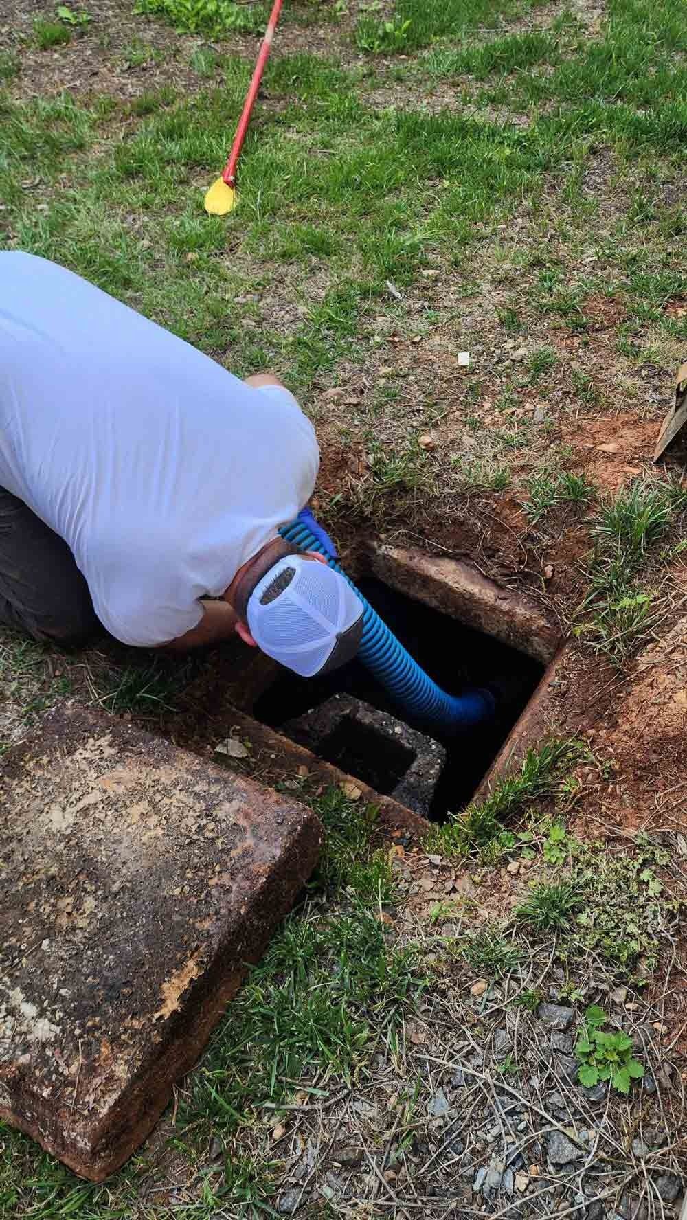 A person leaning into an open square hole with a blue hose, cleaning a septic tank in a grassy area.