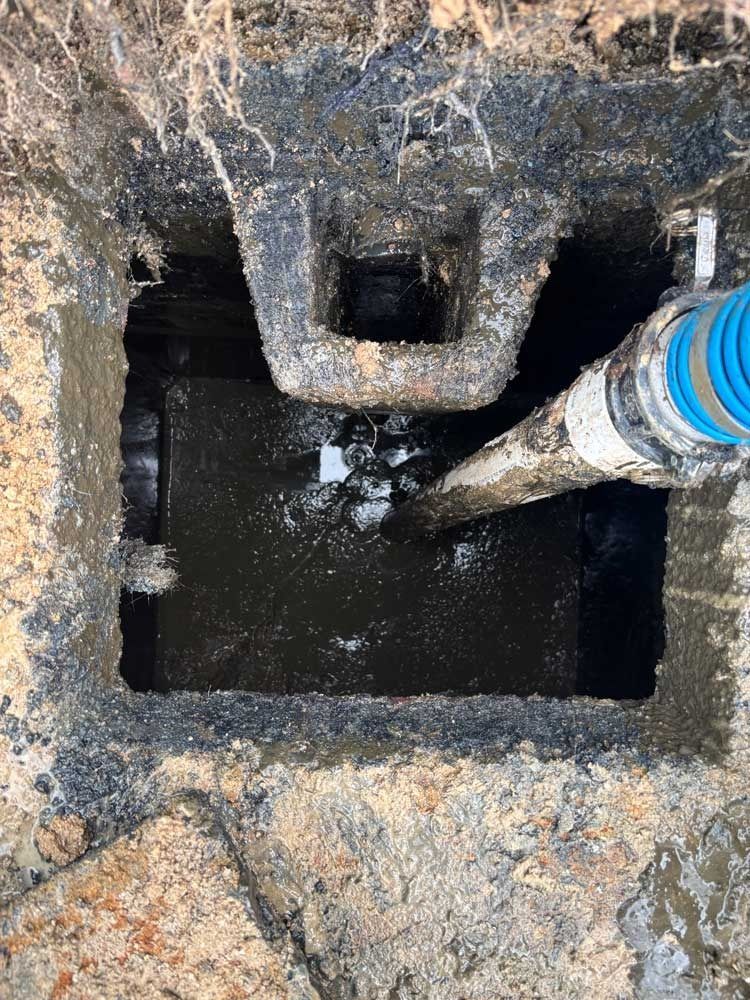 A septic tank being pumped out. A large square hole with a hose spraying water. Dark liquid and mud visible.