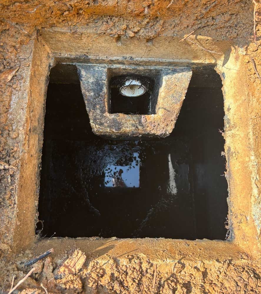 Open, square-shaped access hole in dirt with dark liquid inside; a concrete brace structure is above the liquid.