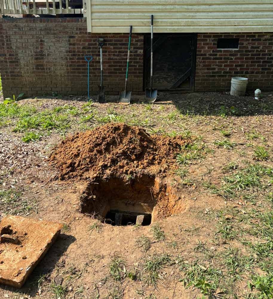 A dug hole in the yard with tools, a dirt pile, and a building with a door and window in the background.