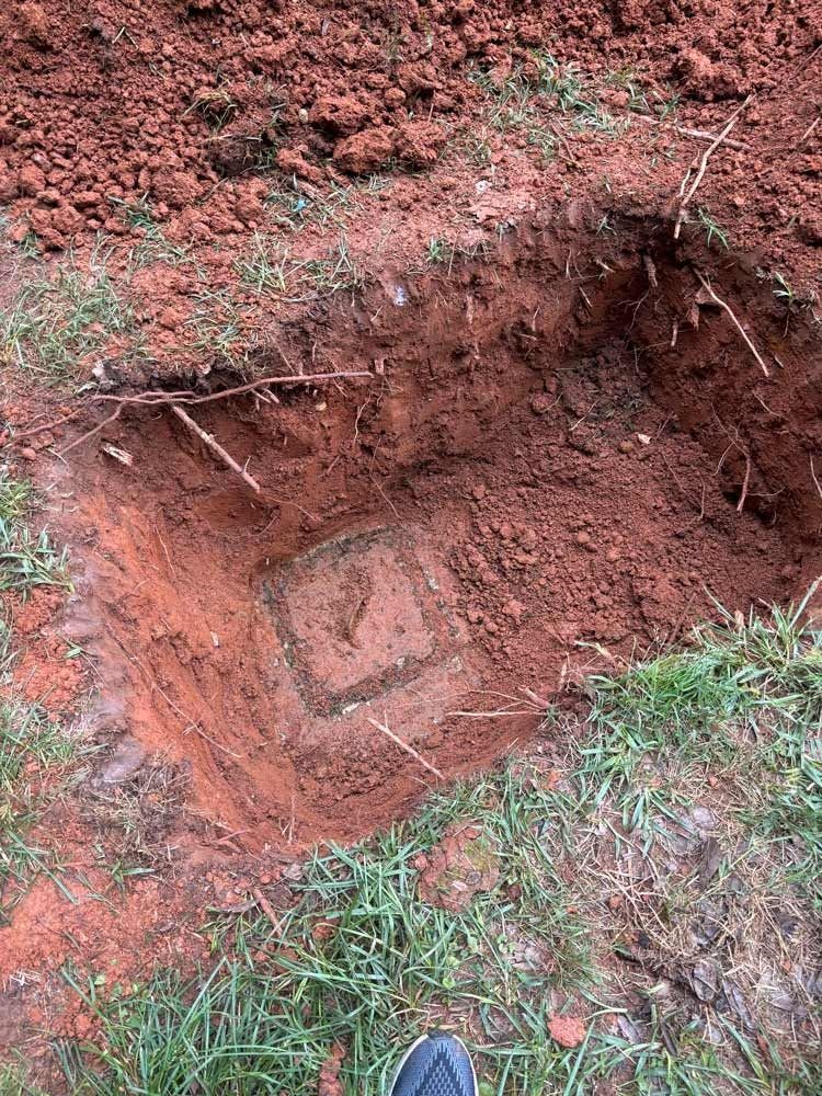 Square hole dug in red soil, revealing a concrete structure. Grass surrounds the hole.