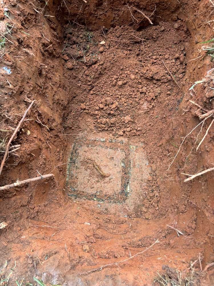 A square hole in reddish-brown earth reveals a concrete marker with weathered markings.
